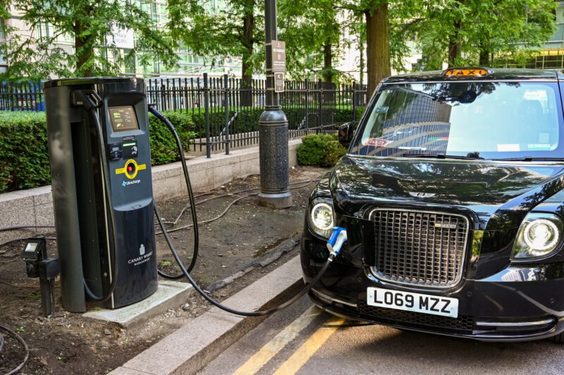Electric black taxi cab parked up in front of a public park, plugged into charging station.