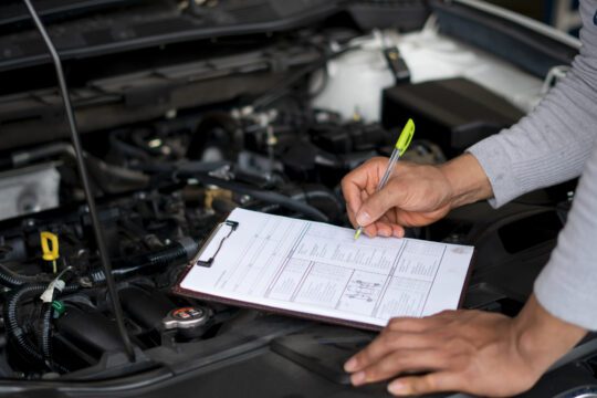 Auto mechanic (or technician) writing on clipboard and checking car engine at the garage