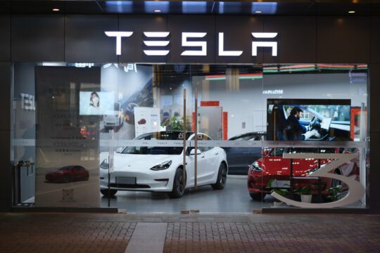 Exterior view of a Tesla showroom at night, featuring two Tesla cars prominently displayed inside. A white Tesla Model 3 is in the foreground, positioned near large glass windows, with a red Tesla car visible behind it. The showroom is well-lit, with a modern interior and a large, illuminated Tesla logo above the entrance. A digital screen inside the showroom displays visuals of Tesla vehicles, and posters with pricing and model details are visible.