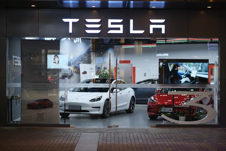 Exterior view of a Tesla showroom at night, featuring two Tesla cars prominently displayed inside. A white Tesla Model 3 is in the foreground, positioned near large glass windows, with a red Tesla car visible behind it. The showroom is well-lit, with a modern interior and a large, illuminated Tesla logo above the entrance. A digital screen inside the showroom displays visuals of Tesla vehicles, and posters with pricing and model details are visible.