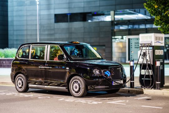 An electric black cab plugged into a charging station on a city street at night, with a modern urban building in the background.