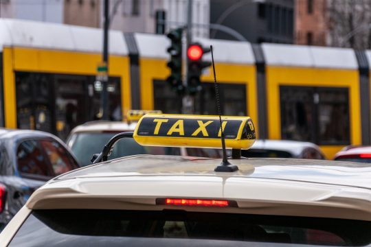 Close-up of a taxi sign on the roof of a vehicle, with bold yellow and black lettering. In the background, a yellow tram is passing by, and a red traffic light is visible, indicating stopped traffic.