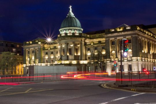 The Mitchell Library in Glasgow city centre, a historic building with a domed roof, is illuminated at night against a deep blue sky. A busy intersection shows red light trails from passing vehicles, captured using a long-exposure technique. A traffic light displaying red, amber, and green signals is visible to the right